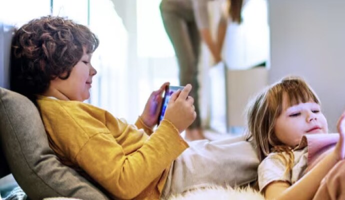 Niños recostados usan tablets mientras la madre en el fondo organiza la cocina.